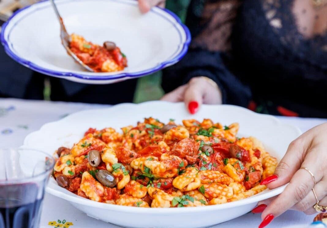 Hands serving pasta with tomato sauce and olives at a table, adorned with patterned cloth and dinnerware. A glass of red wine and a bowl of olives are on the table.