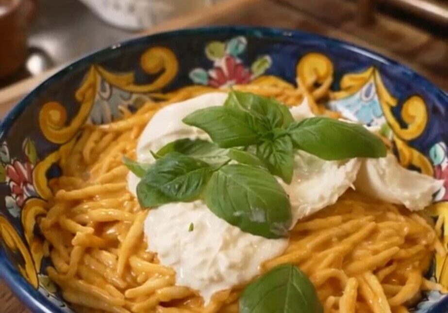 A colorful bowl of spaghetti with red sauce, topped with mozzarella and fresh basil leaves, sits on a wooden counter. A teapot is visible in the background.
