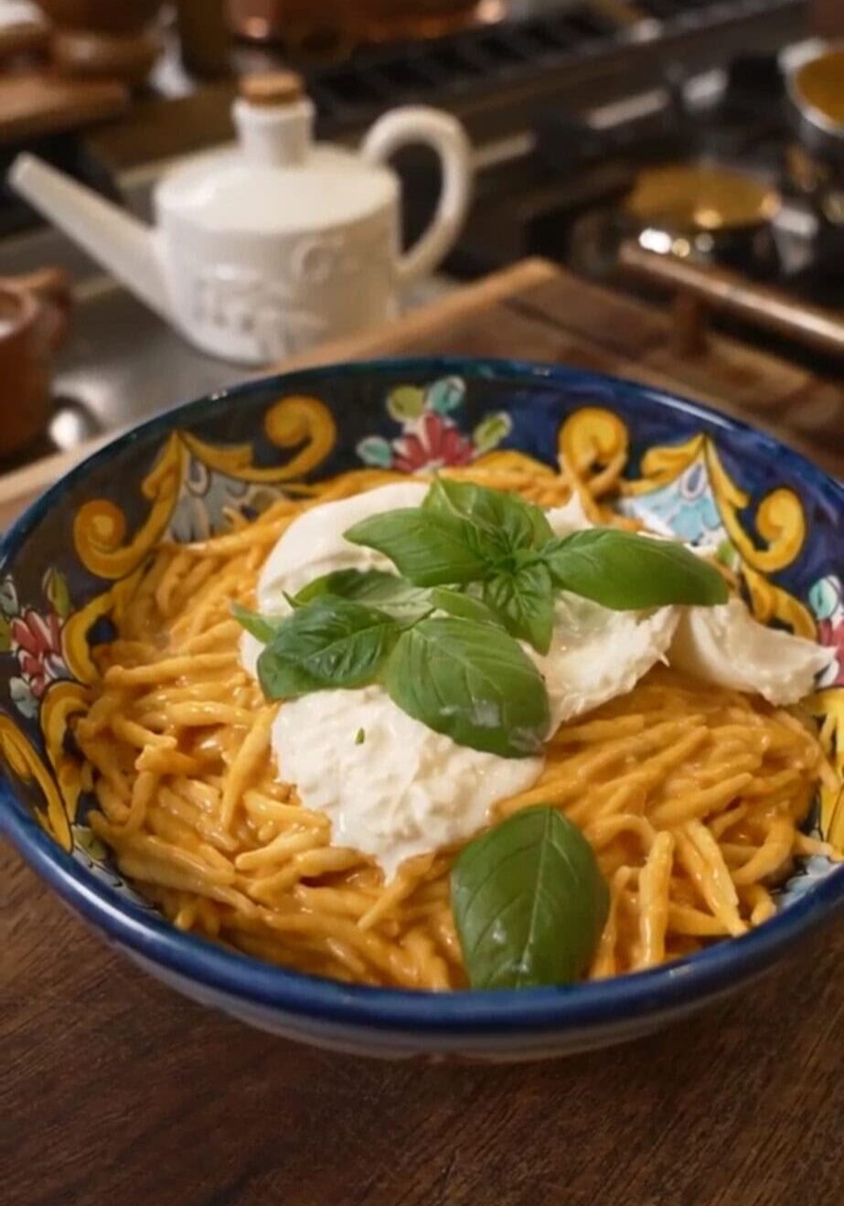 A colorful bowl of spaghetti with red sauce, topped with mozzarella and fresh basil leaves, sits on a wooden counter. A teapot is visible in the background.