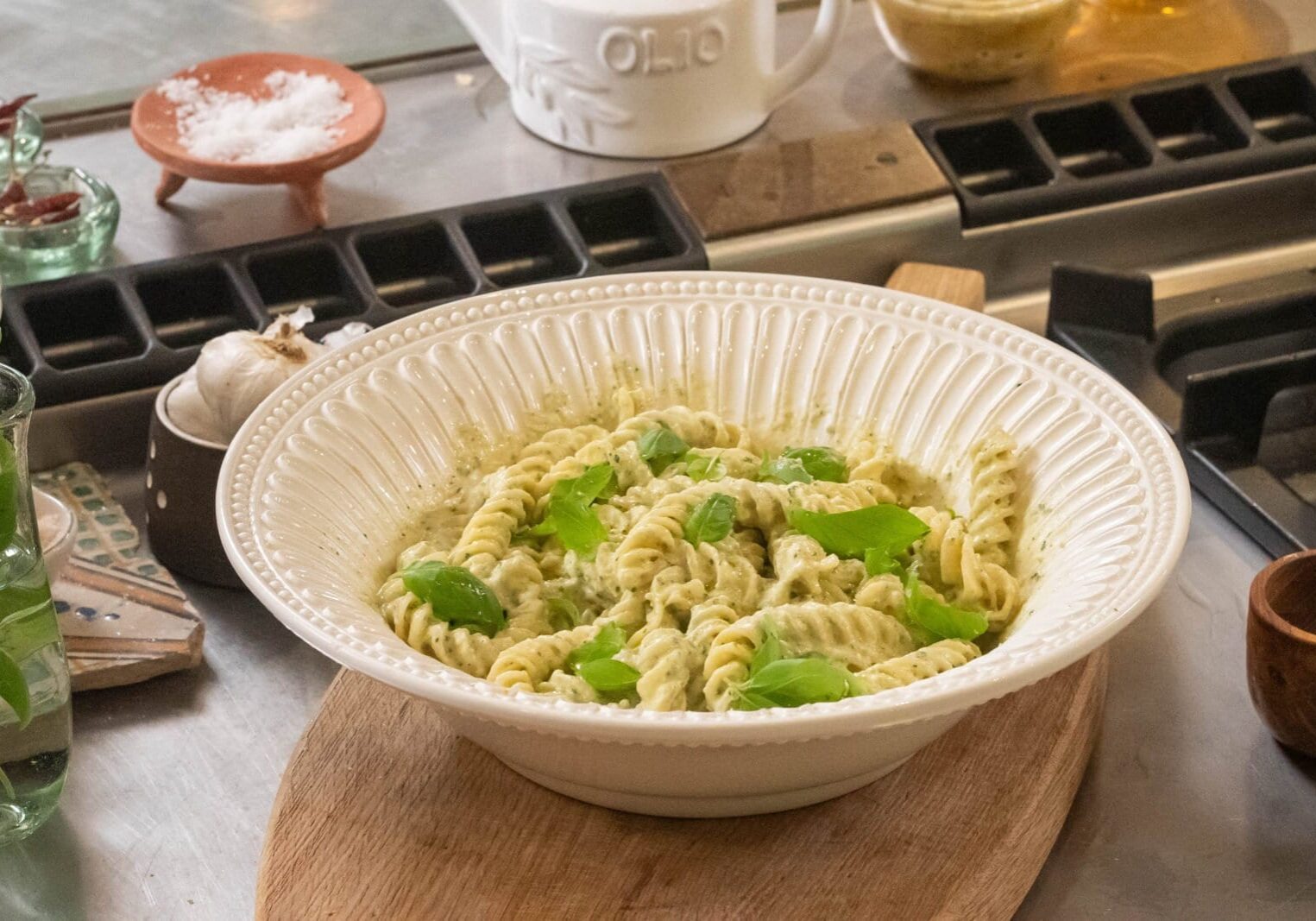A white bowl of fusilli pasta coated in pesto sauce, garnished with fresh basil leaves, placed on a wooden board on a metal countertop with assorted kitchen items in the background.
