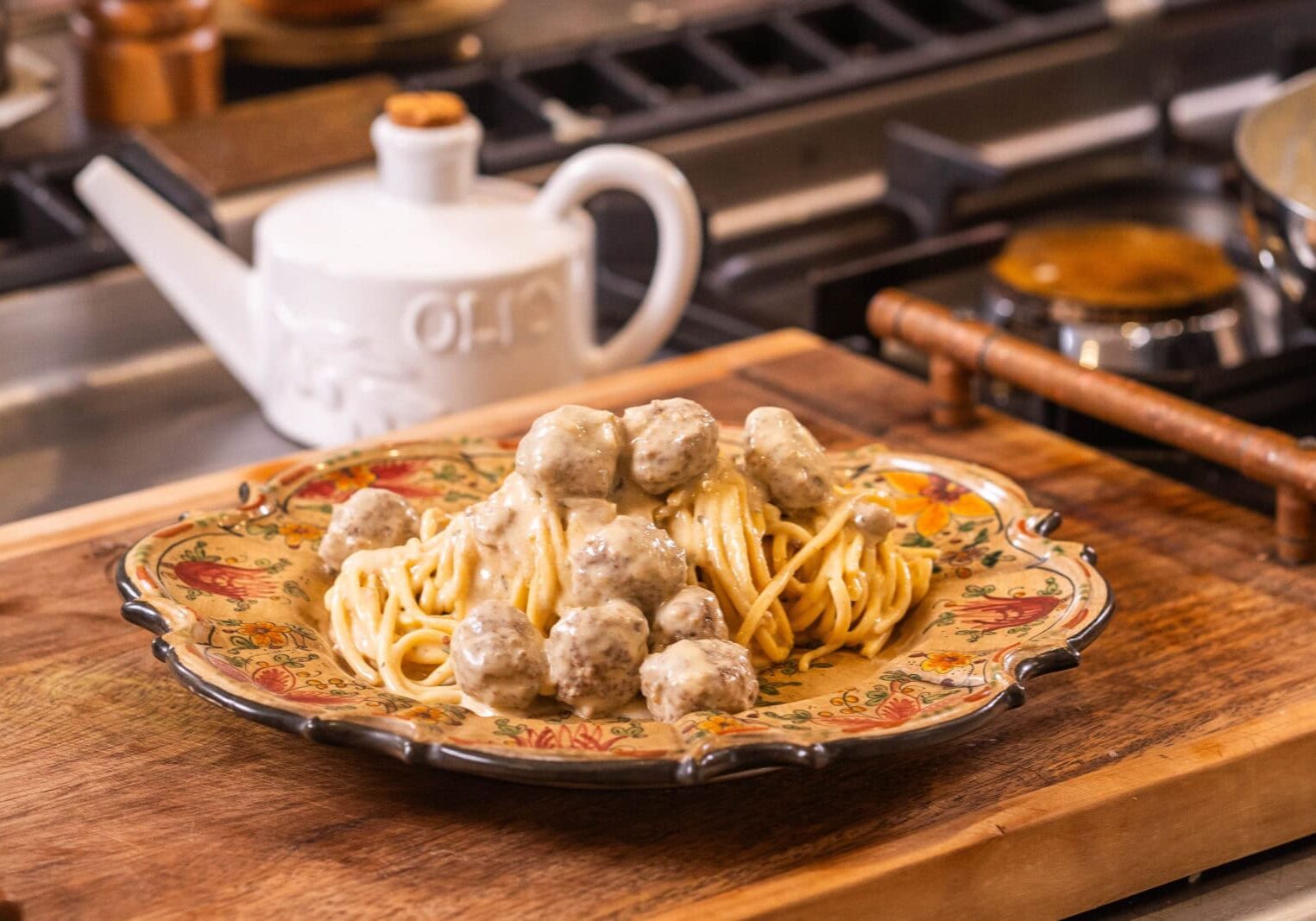 A plate of spaghetti topped with meatballs and creamy sauce on a wooden board, beside a white teapot in a kitchen setting.