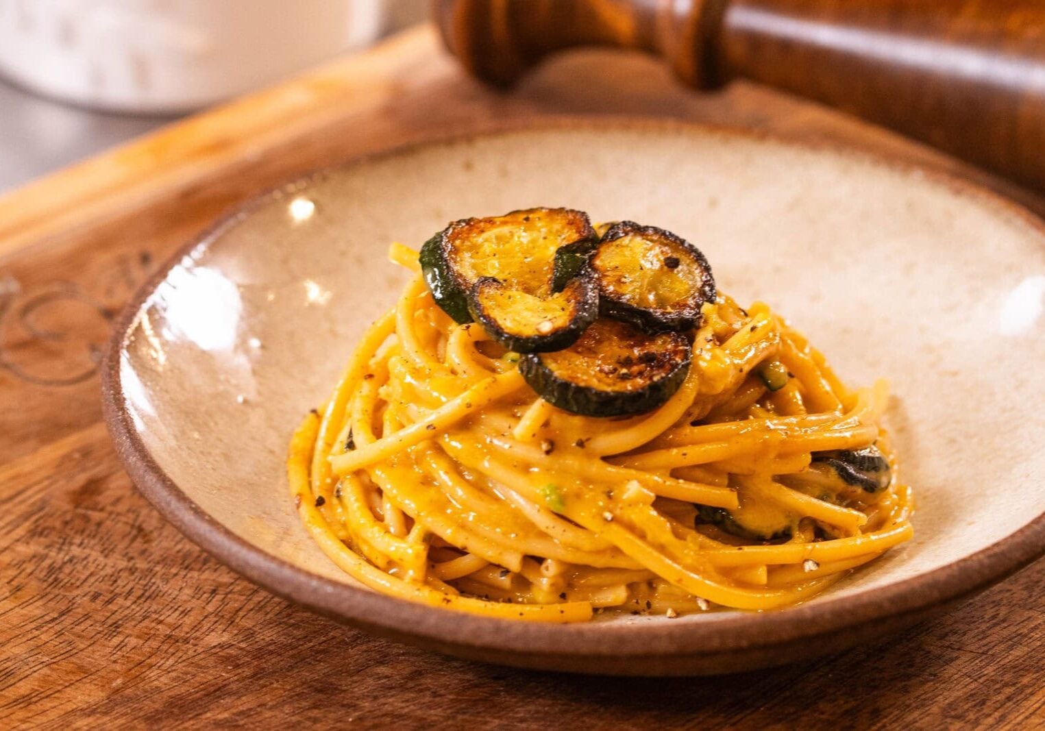 A plate of spaghetti in a creamy sauce topped with grilled zucchini slices, served on a wooden board.