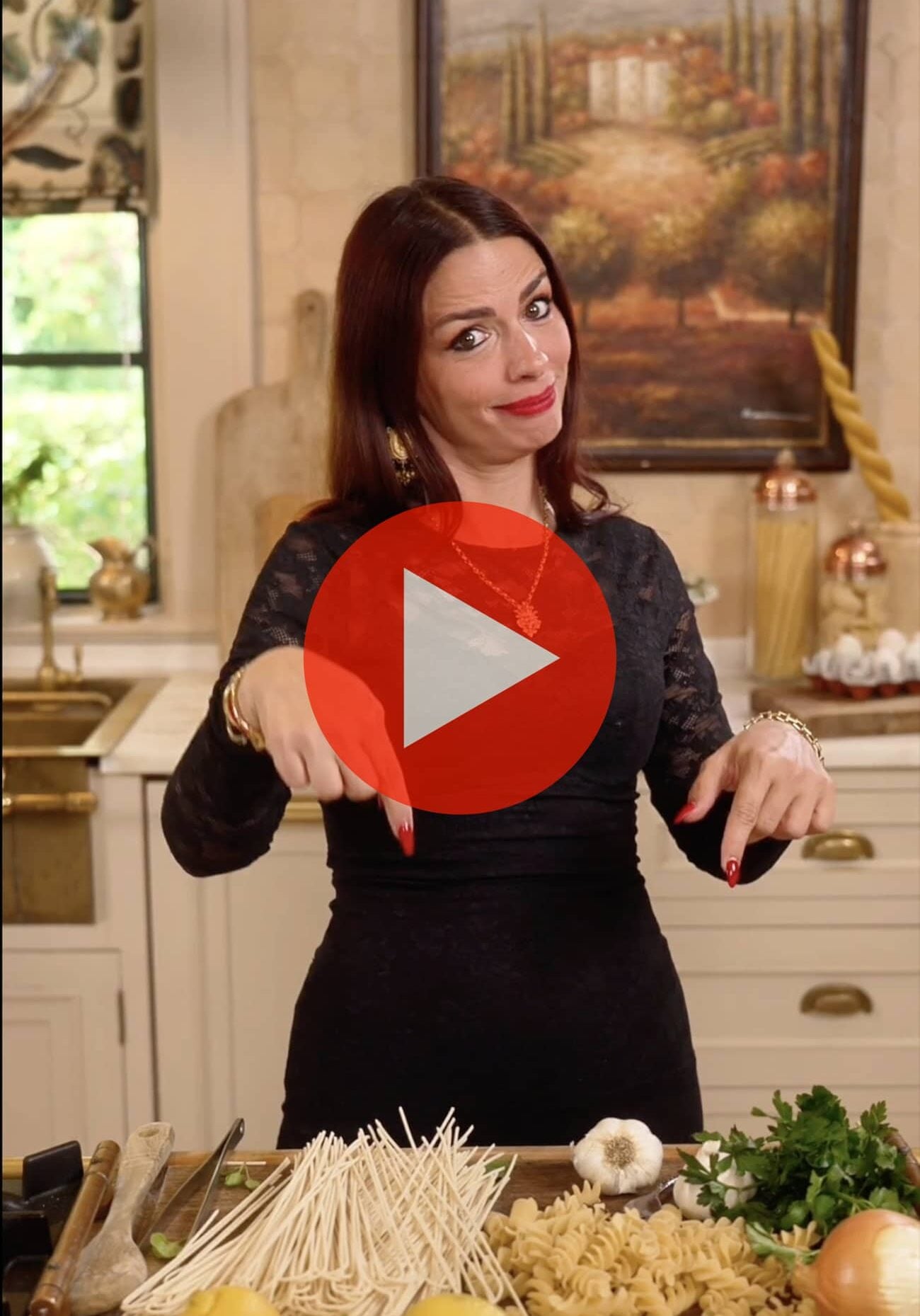 A woman in a black dress stands in a kitchen, pointing down at fresh ingredients on the counter. A large red play button overlays the image.
