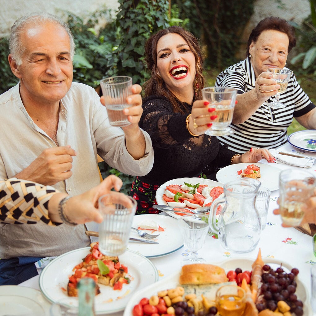 Group of people sitting at an outdoor table, raising glasses and smiling, with plates of food, fruit, and bread in front of them.
