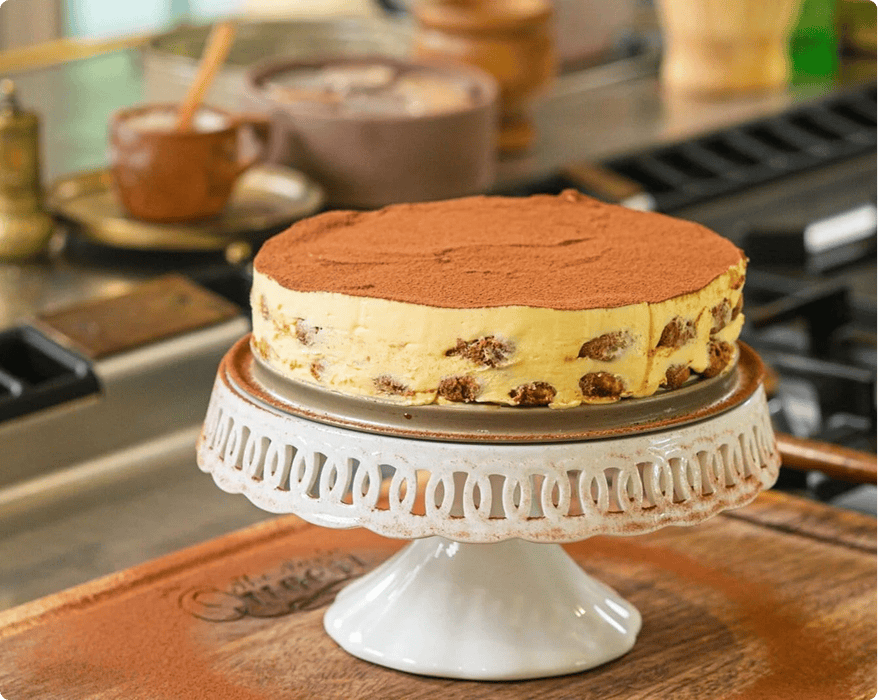 A round tiramisu cake dusted with cocoa powder sits on a white cake stand in a kitchen setting.