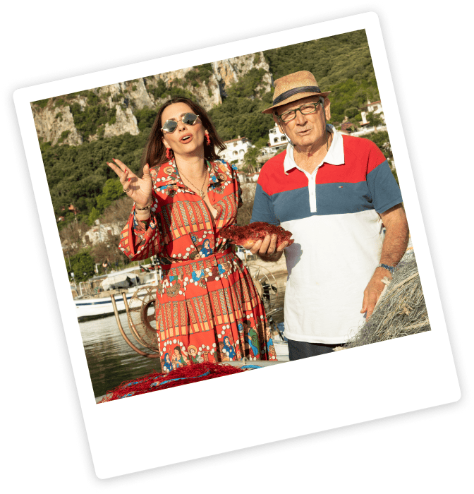 A woman in a patterned dress and sunglasses stands next to an older man in a hat and polo shirt; she holds up a peace sign while he holds a cooked lobster. Outdoor, waterfront setting.