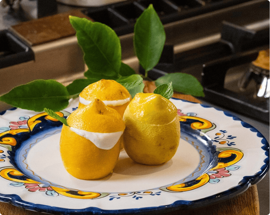 Three hollowed lemons filled with a white cream sit on a decorative plate, garnished with green leaves, on a kitchen stovetop.