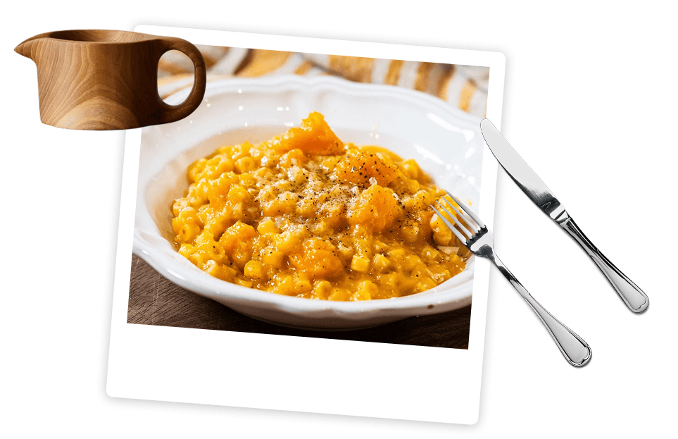 A plate of creamy risotto with pieces of orange squash, next to a wooden sauce pitcher, a fork, and a knife on a white background.