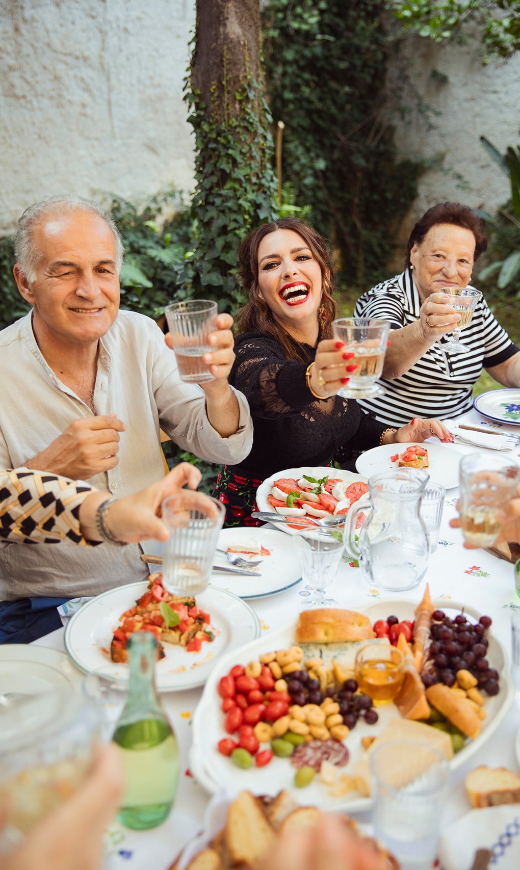 Group of people sitting at an outdoor table, raising glasses and smiling, with plates of food, fruit, and bread in front of them.