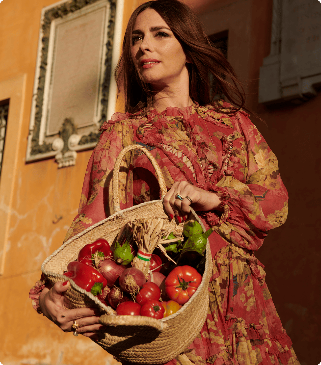 A woman in a floral dress holds a woven basket filled with assorted fresh vegetables, including tomatoes and peppers, standing outdoors near an orange wall.